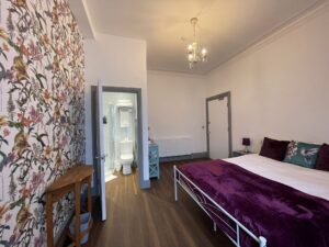 Bedroom with a large double bed, purple throw and floral patterned wallpaper statement wall.