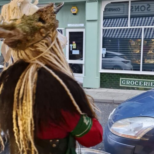 3fe784f8-a829-41a0-a682-cdbc1e8052df A person stood looking across the road, wearing an outfit for the Krampus Run, and holding a drum